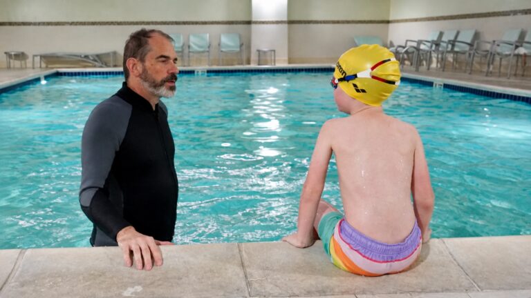 Swim instructor talking with a child wearing a yellow swim cap at the pool edge.